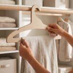 Persona colgando suavemente un pantalón de lino en un gancho de cartón comprimido sin pinzas dentro de un clóset iluminado.